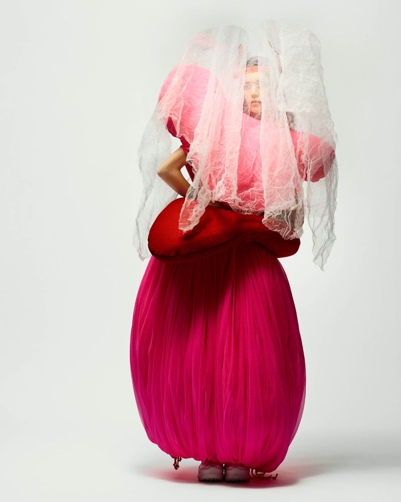 Fashion portrait of a woman in a pink sculptural tulle dress and a white lace veil, posing in a bright white photo studio.
