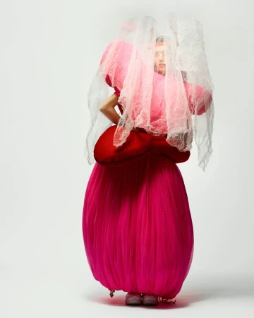 Fashion portrait of a woman in a pink sculptural tulle dress and a white lace veil, posing in a bright white photo studio.