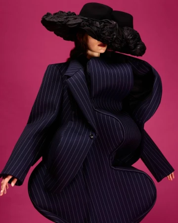 Fashionable adult posing in an oversized pinstripe suit and black hat with ruffles against a vivid pink backdrop in a studio setting.