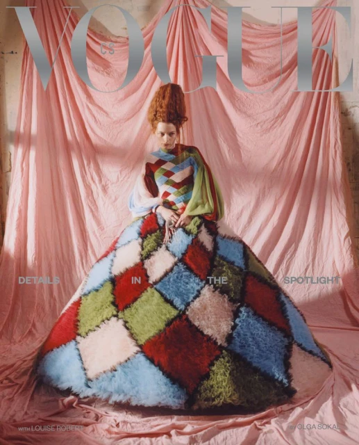 A woman with red hair poses in a colorful patchwork dress against a pink backdrop for a Vogue magazine cover shoot in a photo studio.