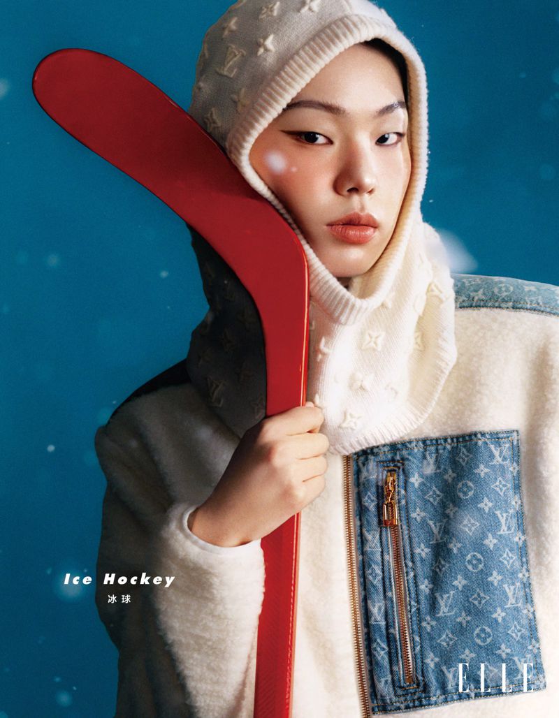 Portrait of an Asian woman in a designer outfit holding a red ice hockey stick against a blue background in a studio setting.