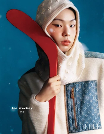 Portrait of an Asian woman in a designer outfit holding a red ice hockey stick against a blue background in a studio setting.