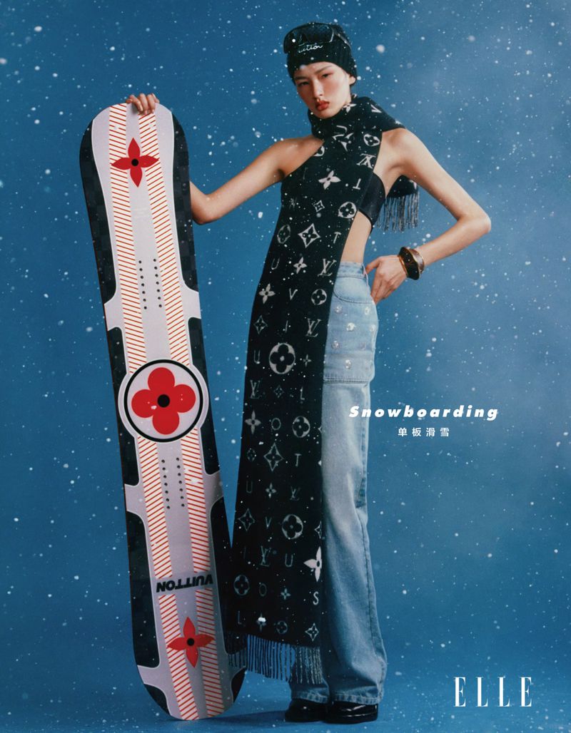 Editorial fashion shot of a woman in winter clothing, holding a snowboard with a logo, against a blue background with snow.