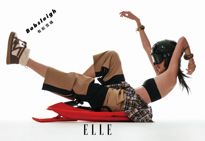 A stylish adult woman with a helmet and ski goggles poses on a red snow sled in a studio with white backdrop, showcasing a fashion and lifestyle scene.