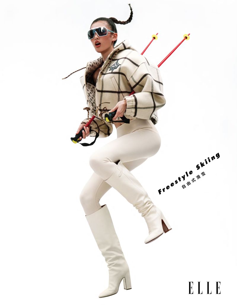 A stylish woman poses in a ski outfit against a bright background, holding ski poles and wearing sunglasses and heeled boots in a studio shot.