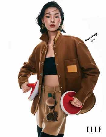A woman poses in a brown jacket, black top, and tan pants, holding curling stones in a studio shot against a white backdrop.