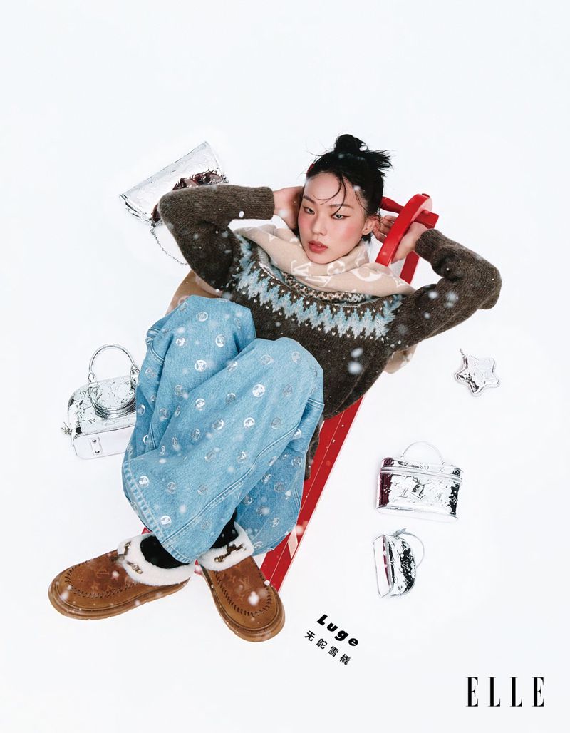A young woman lies on a red sled surrounded by silver handbags and wearing wintery clothes in a studio fashion editorial.