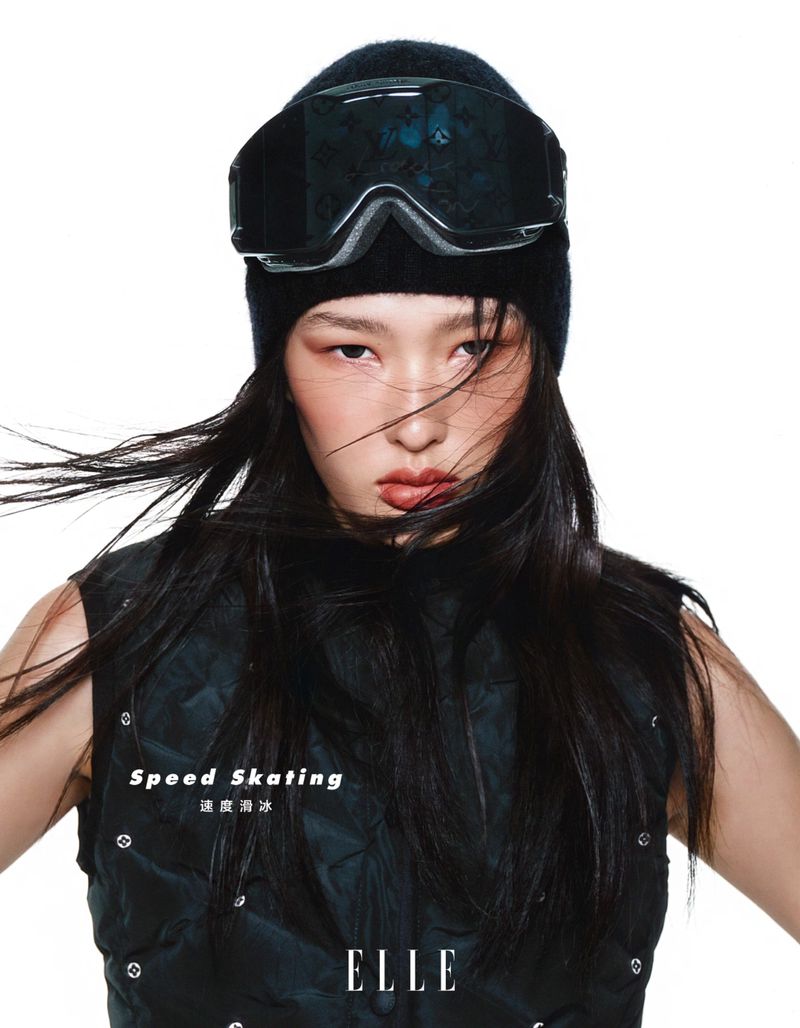 Eye-level portrait of a young woman with black ski goggles and a beanie, wearing a dark puffy vest against a white background.