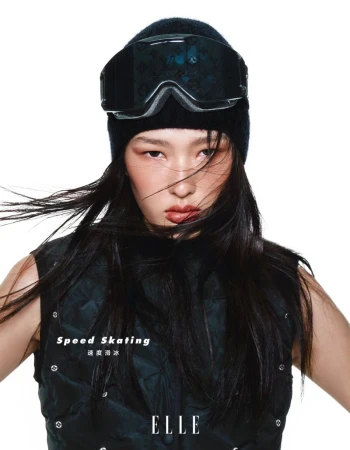 Eye-level portrait of a young woman with black ski goggles and a beanie, wearing a dark puffy vest against a white background.