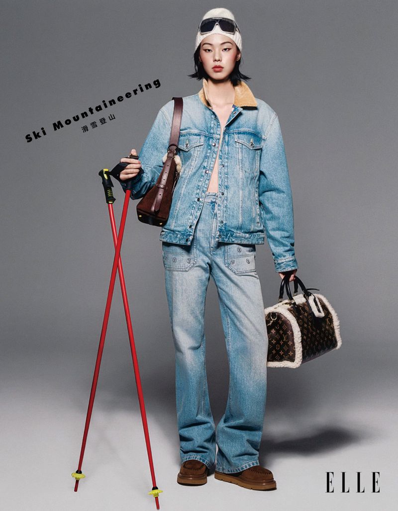 A woman in ski gear with a denim jacket, ski goggles and a beanie hat stands in a studio, holding ski poles and a fur bag.