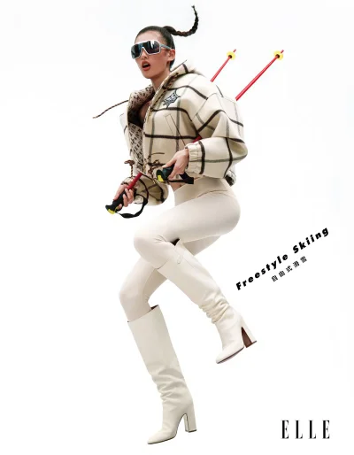 Model in plaid puffer jacket and white boots leaping with ski poles against a white background for Elle China.