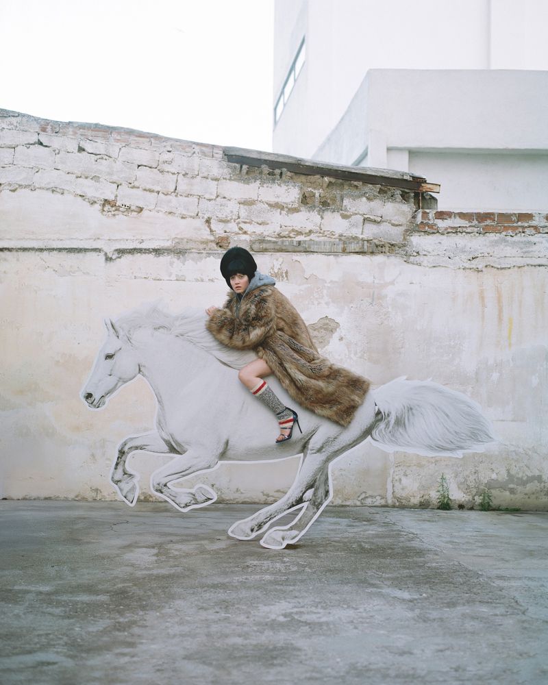 A woman wearing a fur coat and hat sits atop a stylized white horse, posing outdoors with an urban brick wall in the background.