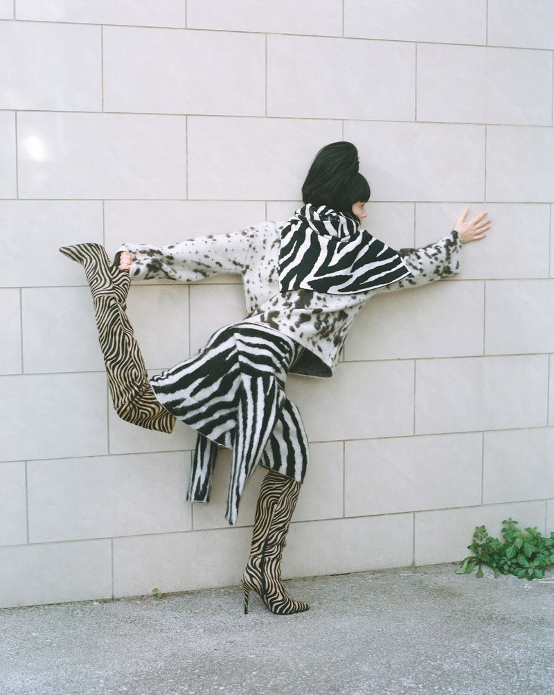 A woman in zebra print poses against a wall, wearing a zebra print jacket, skirt, and knee-high stiletto boots; fashion shot.