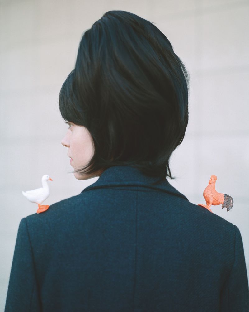 A woman with a dark blue coat and dark hair stands in front of a plain wall with toy bird figurines on her shoulders.