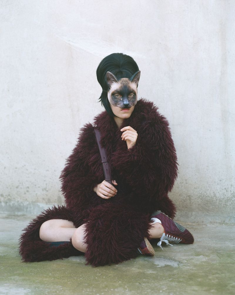 Person in fur coat and animal mask sitting against textured wall, holding a dark object in their hands, surreal fashion concept.