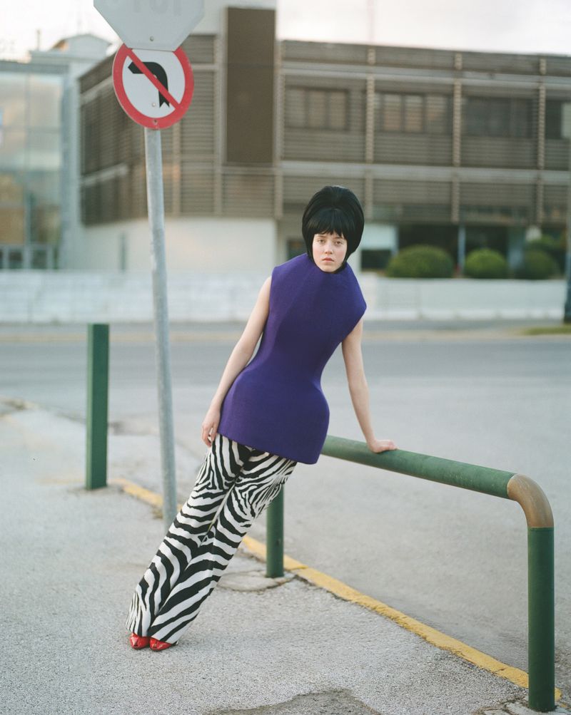Young woman in vibrant blue dress, zebra pants, red shoes leaning on a green rail against an urban background with a traffic sign.