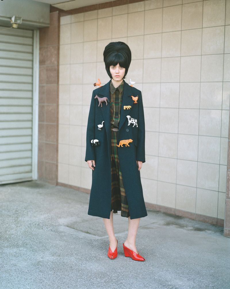 A fashionable young woman with a striking beehive hairstyle poses outdoors in a long coat adorned with various animal brooches.