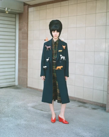 A fashionable young woman with a striking beehive hairstyle poses outdoors in a long coat adorned with various animal brooches.