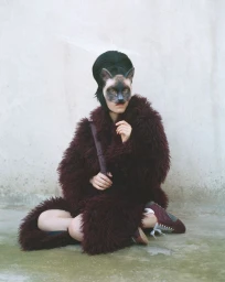 Model wearing a cat mask and fuzzy burgundy coat seated against a textured concrete wall