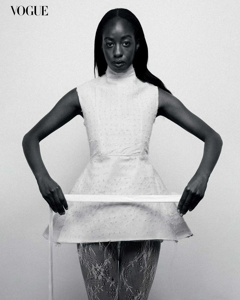 Vogue fashion portrait: A Black woman poses indoors wearing a white top and floral-patterned tights, holding a white measuring tape.