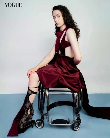 Fashion editorial shot of a young woman in a burgundy dress seated in a wheelchair, wearing a lace boot, and posing indoors.