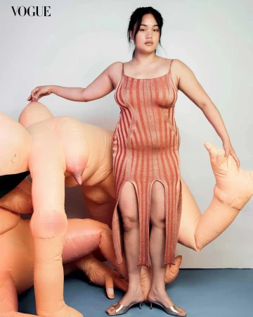 Adult woman posing in vertical striped dress with thigh slits, silver heels, standing next to inflatable human-like form in photo studio.