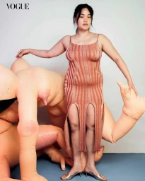 Model standing in an orange ribbed knit midi dress beside large flesh-toned inflatable limbs in a studio.