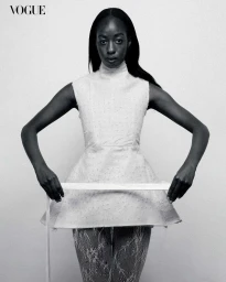 Black female model posing in a high-neck structured white tunic dress and lace tights against a plain studio backdrop.