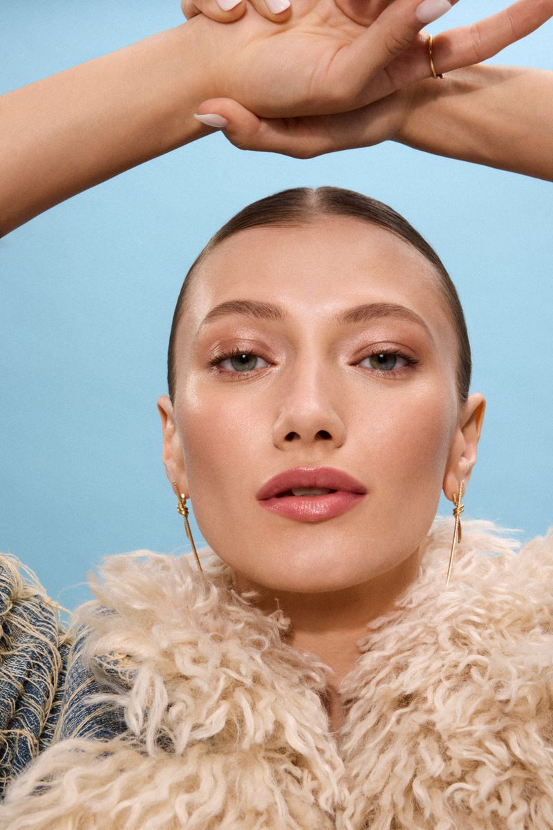 Close-up portrait of a fair-skinned woman with slicked-back hair wearing gold earrings and a fuzzy, textured jacket against a blue background.