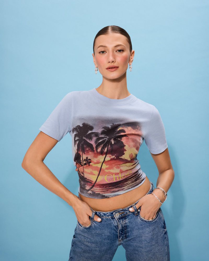A young woman poses against a bright blue backdrop wearing a light blue t-shirt with a beach sunset graphic print and blue jeans.