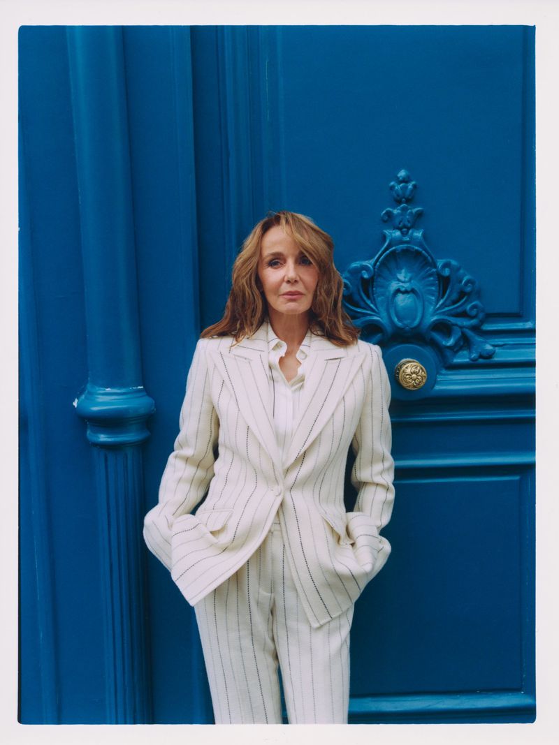 Woman in striped suit poses against a blue door with ornate detailing. Fashion shot captures elegance in an outdoor setting.