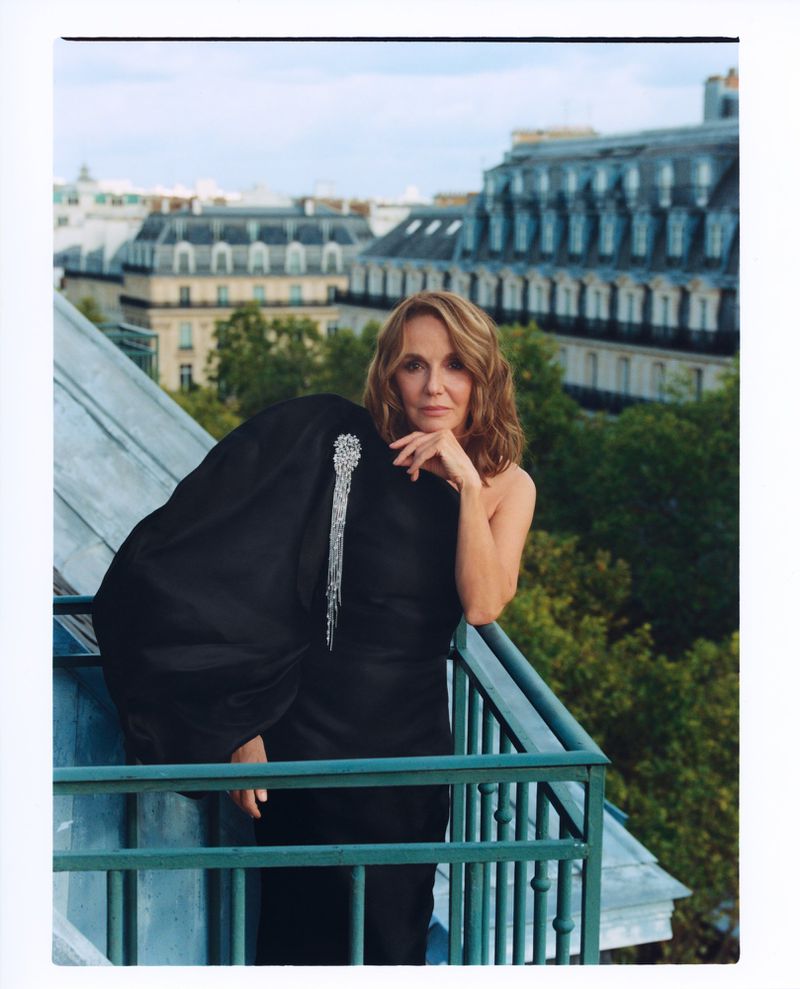 Adult woman poses on a Paris balcony wearing black dress and holding a jacket with silver brooch, overlooking city rooftops.