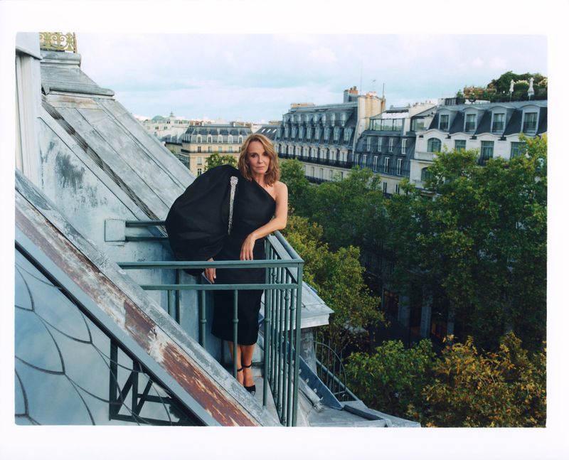 A fashionable adult woman poses in a black dress on a rooftop balcony in an urban environment with city buildings in the background.
