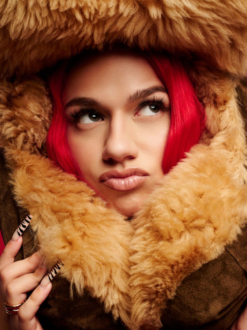 Portrait of a woman with red hair wearing a fur-lined hood, looking upward, with painted fingernails and lip gloss.