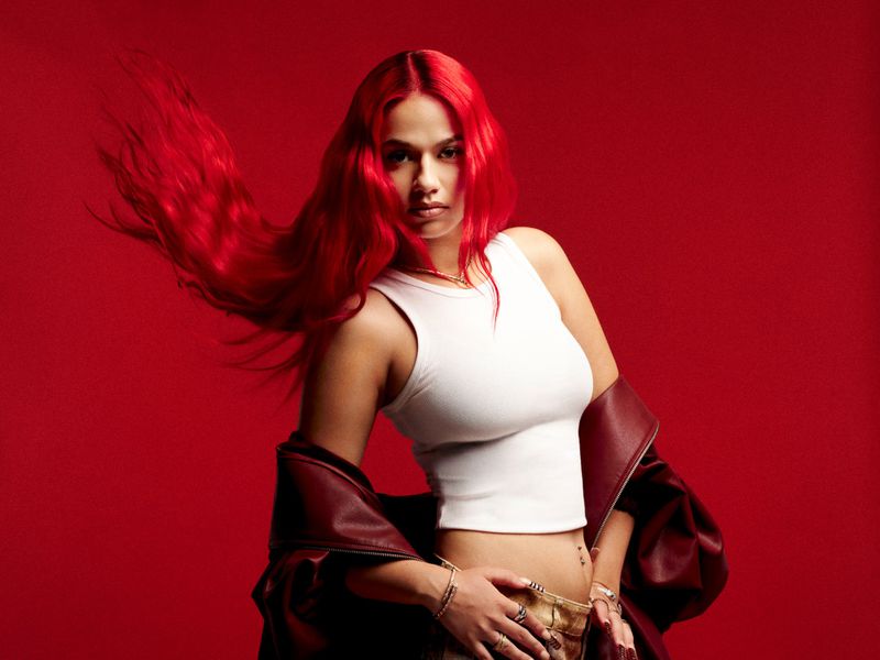 Young woman with long, flowing red hair poses in front of a red background, wearing a white tank top and a red leather jacket.