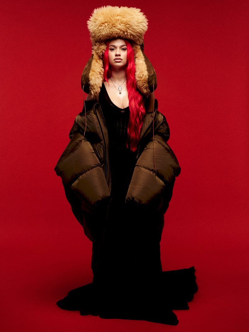 A woman in a large fur hat and oversized puffer jacket poses against a red background, showcasing a blend of fashion and unique style.