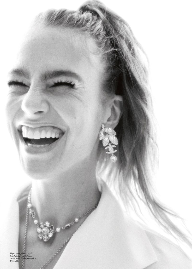 Close-up black and white portrait of a smiling adult female with hair in a ponytail wearing Chanel jewelry against a bright background.