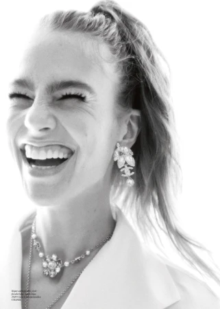 Close-up black and white portrait of a smiling adult female with hair in a ponytail wearing Chanel jewelry against a bright background.