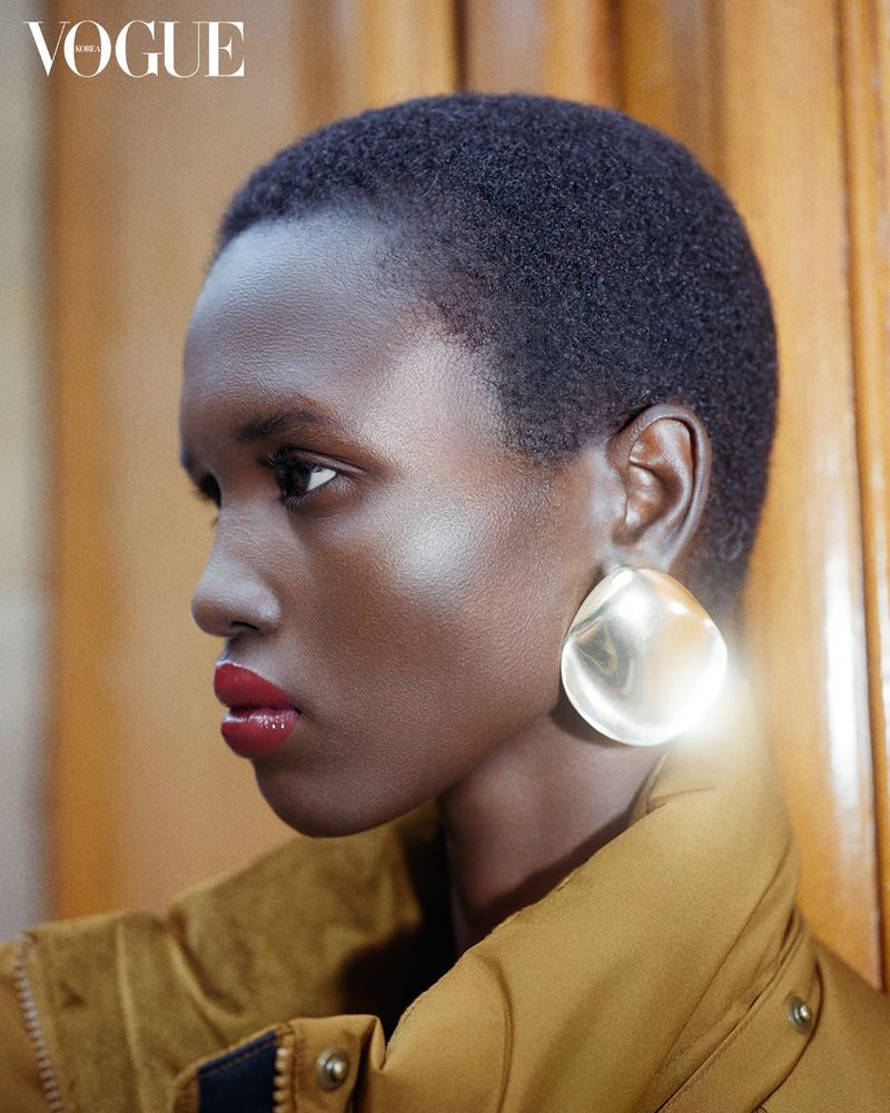 Profile shot of an African American woman with short hair, wearing gold earrings and a brown jacket, against a wood panel backdrop.