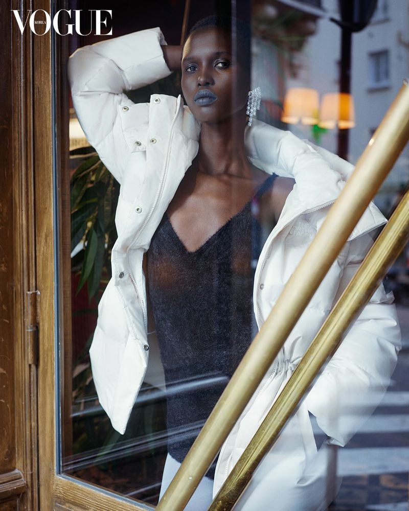 Indoor fashion shot of a dark-skinned woman in a white puffer jacket and dark top, wearing chandelier earrings, in front of a window.