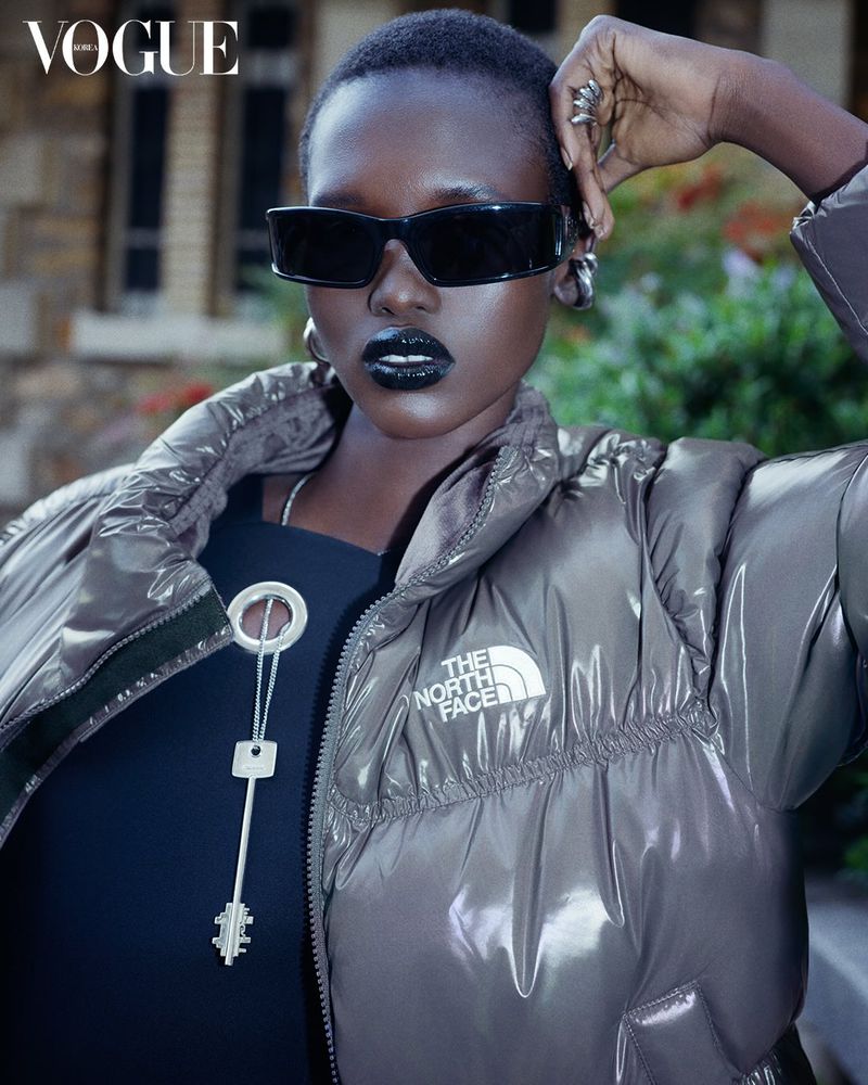 Fashion editorial shot of a stylish African woman wearing a puffer jacket, sunglasses, and bold lipstick, with Vogue logo in the corner.