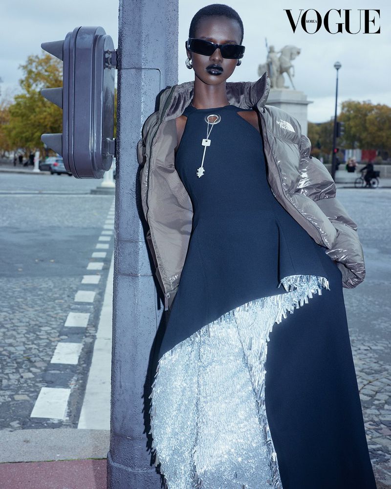 A black woman stands in a street wearing a long dress with fringe detail and a puffer jacket with a traffic light to her side.