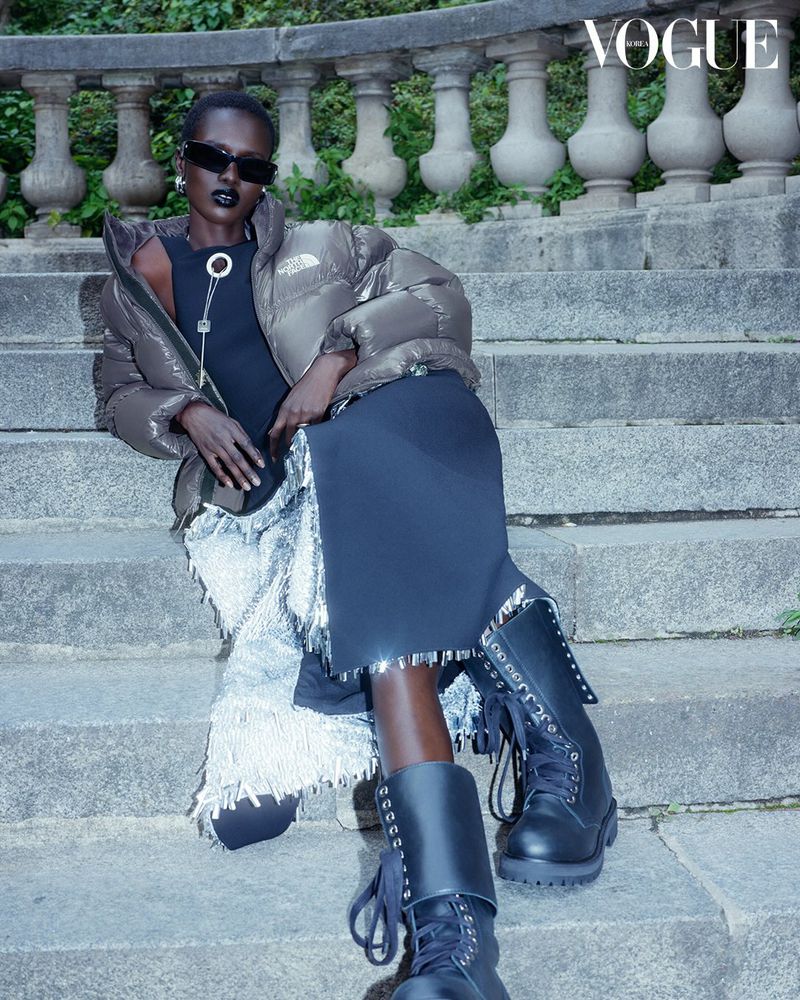 A woman in a puffer jacket and dark lipstick sits on stone stairs with a Vogue logo visible, showcasing fashion and editorial style.