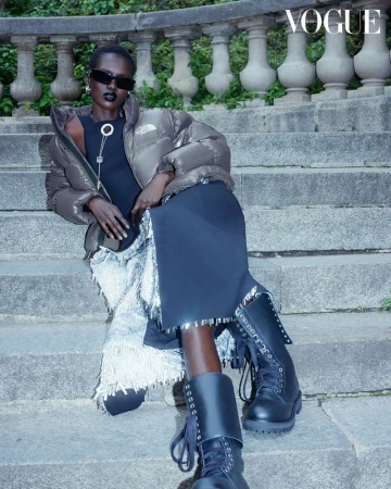 Amar Akway seated on stone steps wearing a taupe puffer jacket, black dress, sunglasses, and combat boots for Vogue Korea.