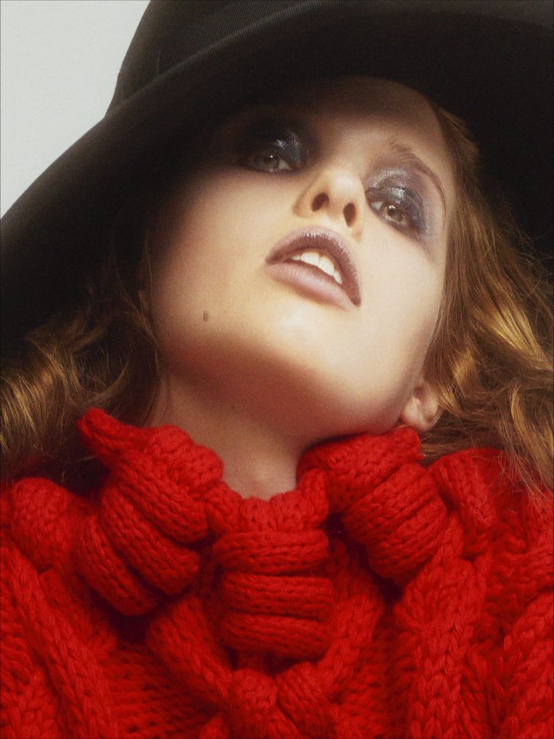 Ever Anderson wearing a red knit sweater and a wide black hat against a neutral backdrop in a portrait by Willy Vanderperre.