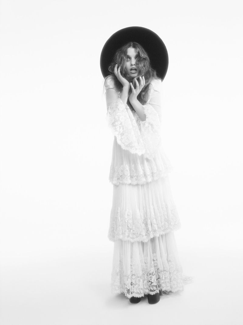 Black and white portrait of a young woman in a tiered lace dress and a wide-brimmed hat, posing with hands near her face on a white backdrop.