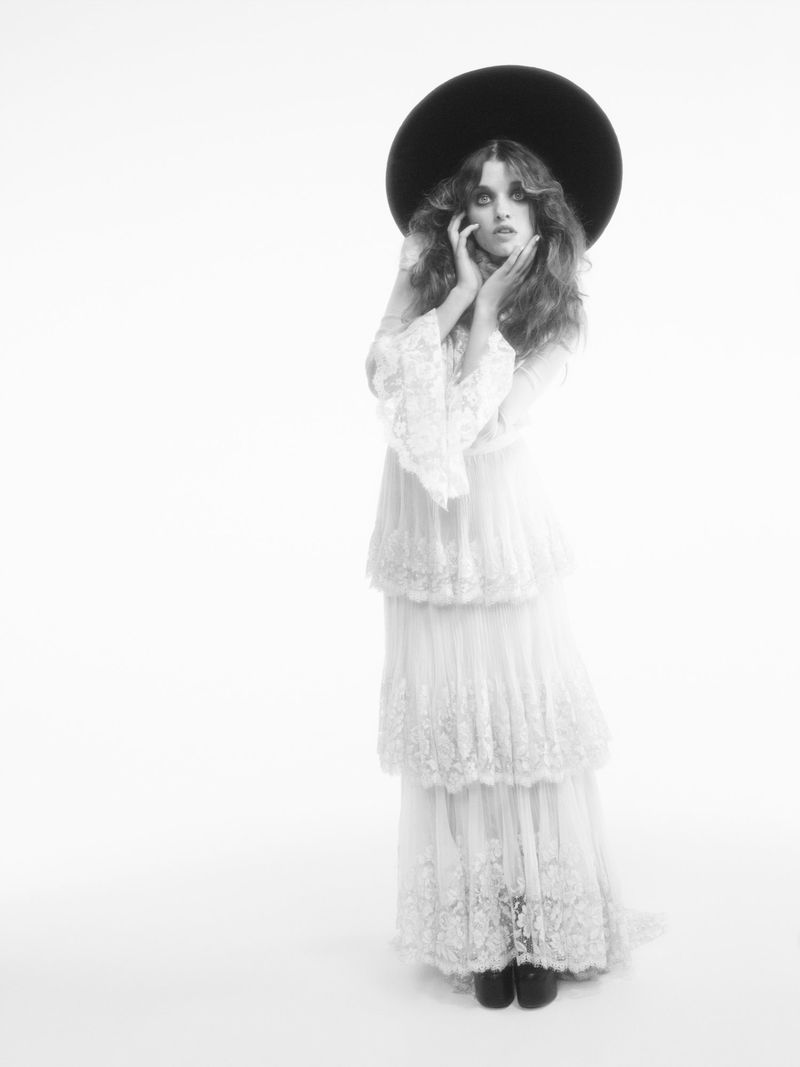 Ever Anderson posing in a full-length tiered white lace dress and oversized black saucer hat against a white seamless background.