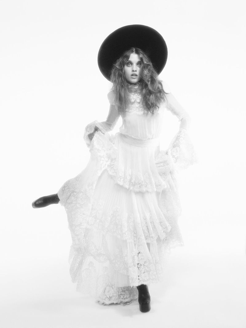 Ever Anderson poses dynamically in a tiered white lace dress and an oversized black wide-brim hat against a white background.