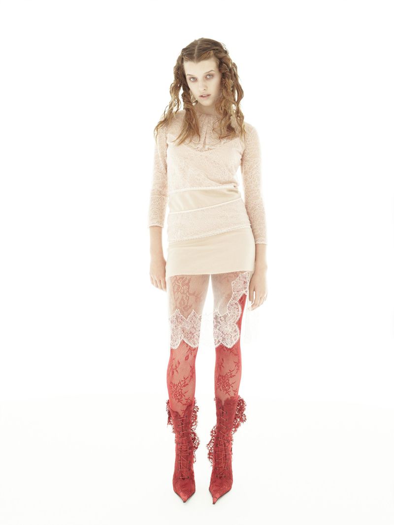 A woman with curly brown hair models a beige dress with lace-trimmed leggings and red lace boots in a brightly lit photo studio.