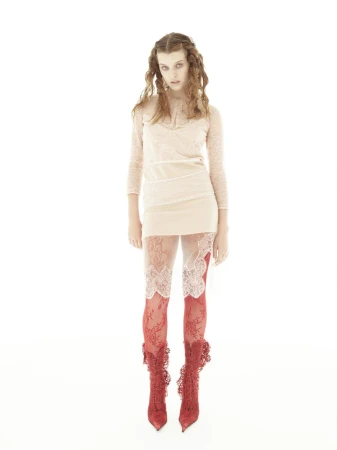 A woman with curly brown hair models a beige dress with lace-trimmed leggings and red lace boots in a brightly lit photo studio.